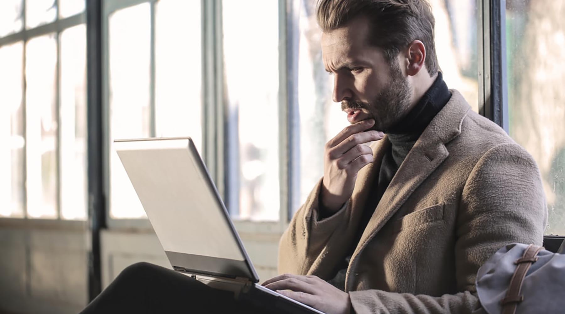 confused looking man staring at laptop with hand on chin wearing brown fuzzy jacket
