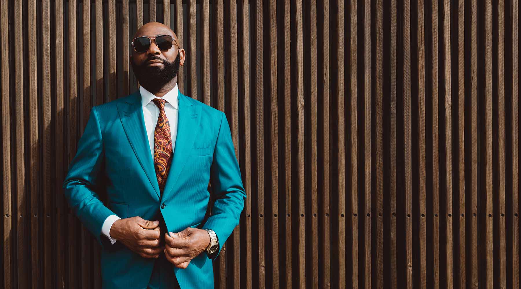 handsome black man in teal suit with orange paisley tie and wooden slat wall background